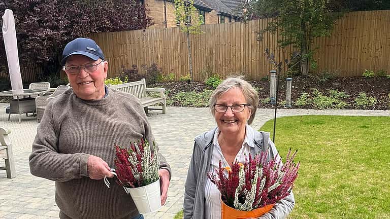 Two people stand outdoors smiling, one holding gardening tools and the other carrying a bunch of freshly picked flowers.