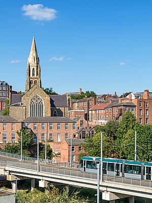 A modern tram gliding through an elevated track, with a backdrop of historic buildings and church towers in Nottingham.