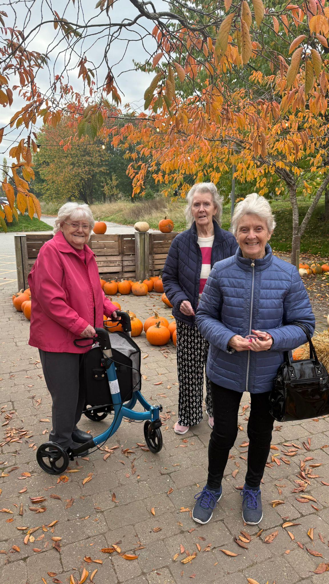 Three older women stand smiling among pumpkins and colourful autumn leaves during a seasonal outing.
