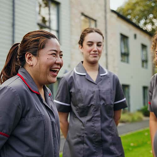 A group of female care professional colleagues in grey uniform conversing and smiling.