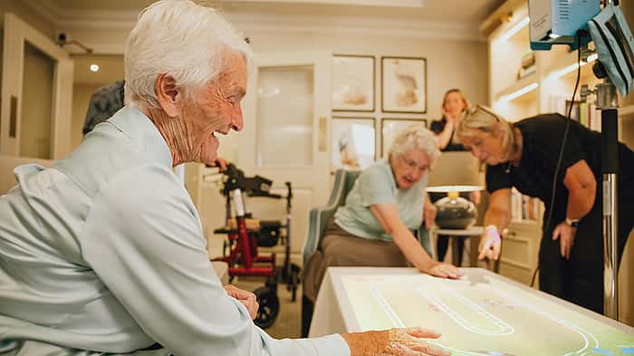 Elderly women interacting with a digital touch table, while others are engaged in the activity in a cozy indoor setting