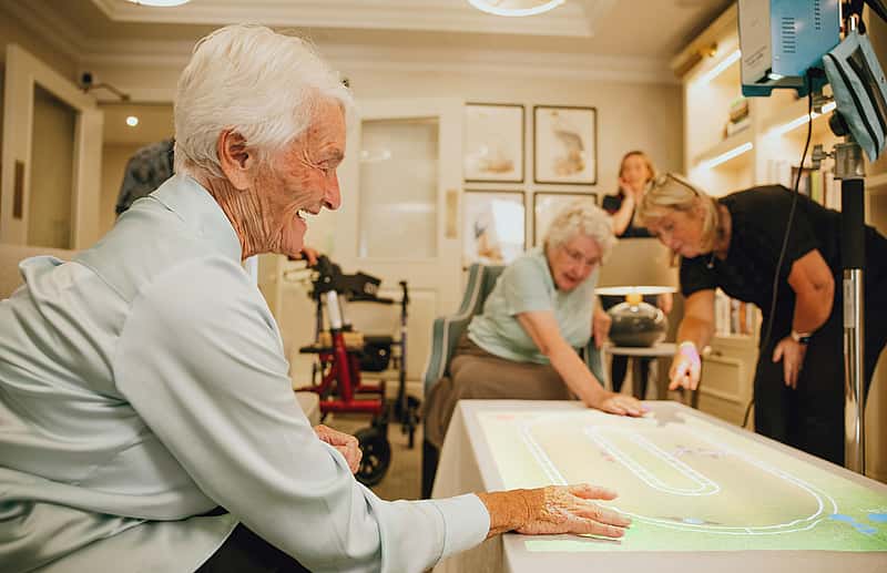 Elderly women interacting with a digital touch table, while others are engaged in the activity in a cozy indoor setting