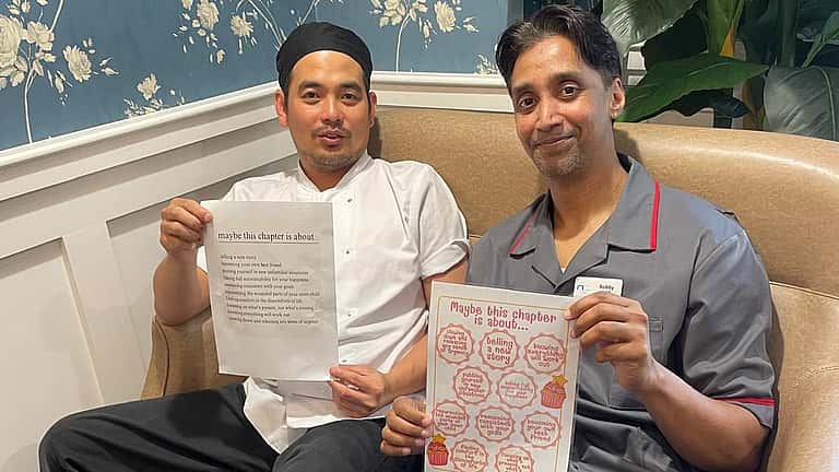 Two men sit on a sofa holding up papers with artwork or designs, smiling proudly at the camera.
