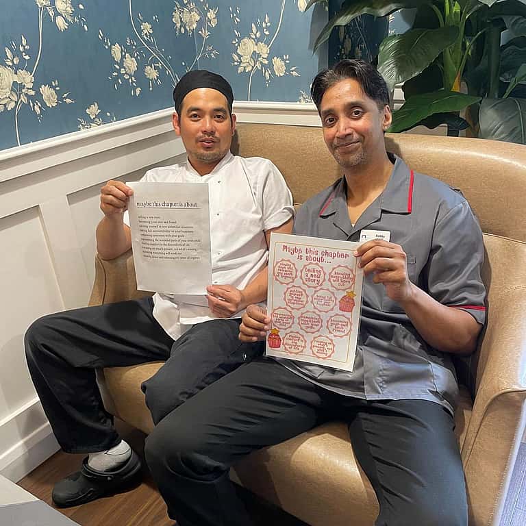 Two men sit on a sofa holding up papers with artwork or designs, smiling proudly at the camera.