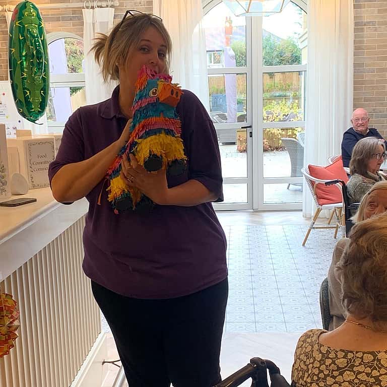 A woman stands indoors holding a brightly colored piñata while others sit nearby.