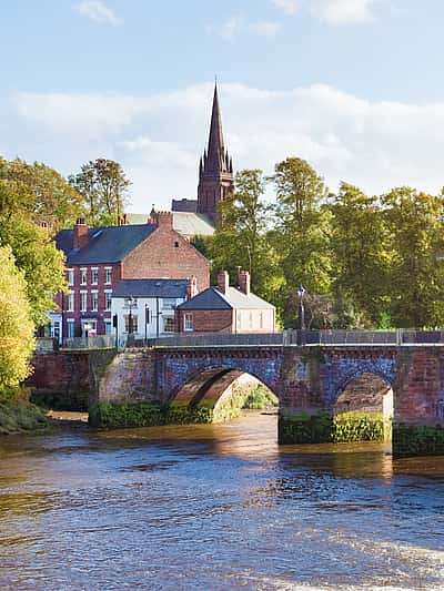 The image captures a charming scene of a stone bridge over a river, with a quaint building and church spire set against a backdrop of lush greenery in Cheshire.
