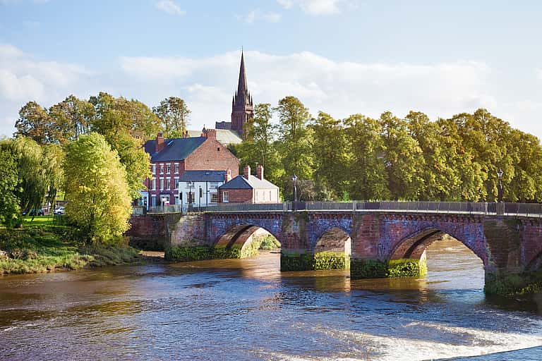 The image captures a charming scene of a stone bridge over a river, with a quaint building and church spire set against a backdrop of lush greenery in Cheshire.