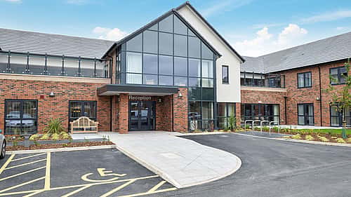 A modern brick-and-glass building with a central reception entrance sits behind a curved walkway and adjacent accessible parking bay.