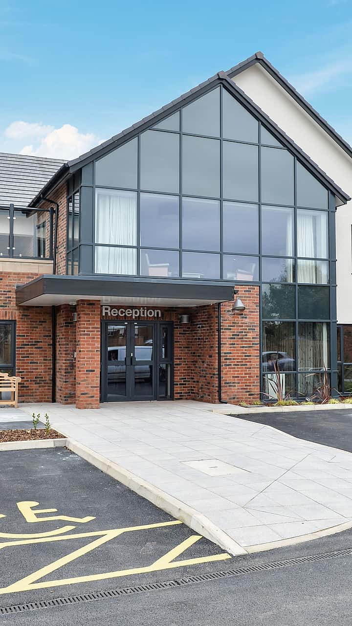 A modern brick-and-glass building with a central reception entrance sits behind a curved walkway and adjacent accessible parking bay.