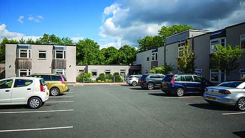 A parking lot in front of a modern, two-story building with several parked cars and greenery in the background under a partly cloudy sky.