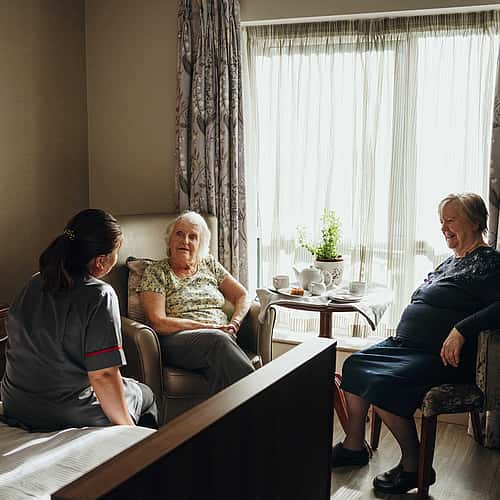 Two elderly women sitting in a cozy room talking to a care professional