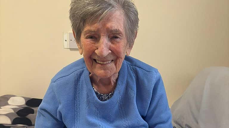 An elderly woman sits smiling with her hands resting on a pink towel, enjoying a gentle pamper or manicure moment.