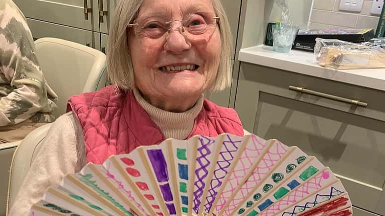 A smiling woman proudly holds a colorful decorative fan spread wide in her hands.