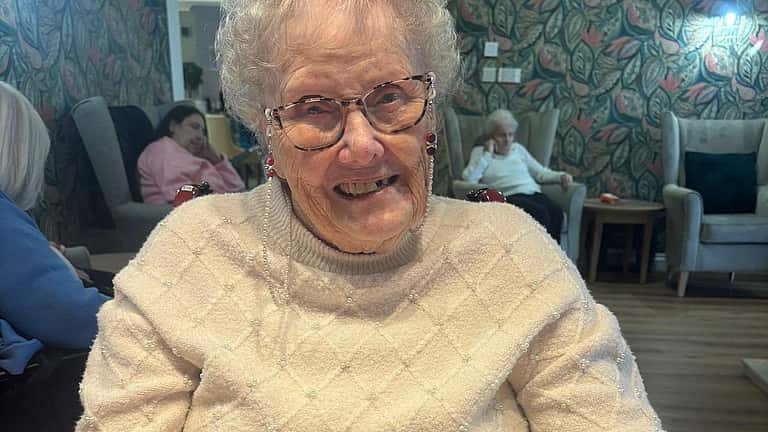 An elderly woman smiles warmly with her hand resting on a pink towel during a relaxing pamper session.
