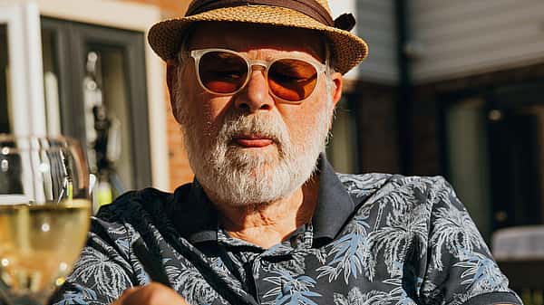 An elderly man wearing a hat and sunglasses is sitting outdoors, writing in a notebook with a glass of wine nearby