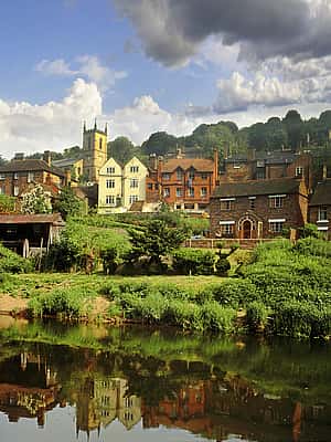 A picturesque view of a village in Shropshire, with colorful houses reflected in a calm river under a dramatic sky.