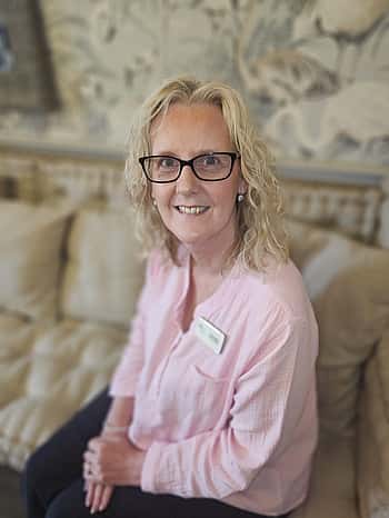 A lady in a pink top with blonde hair and glasses, sat on a sofa.
