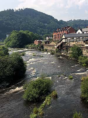 A scenic view of a river flowing through a town in Powys, with lush green hills in the background and a stone building along the riverside.