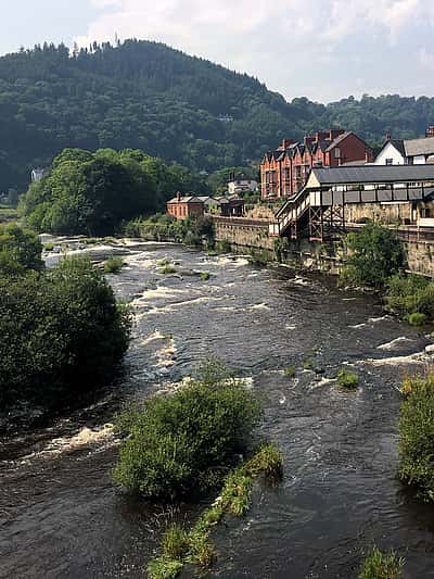 A scenic view of a river flowing through a town in Powys, with lush green hills in the background and a stone building along the riverside.