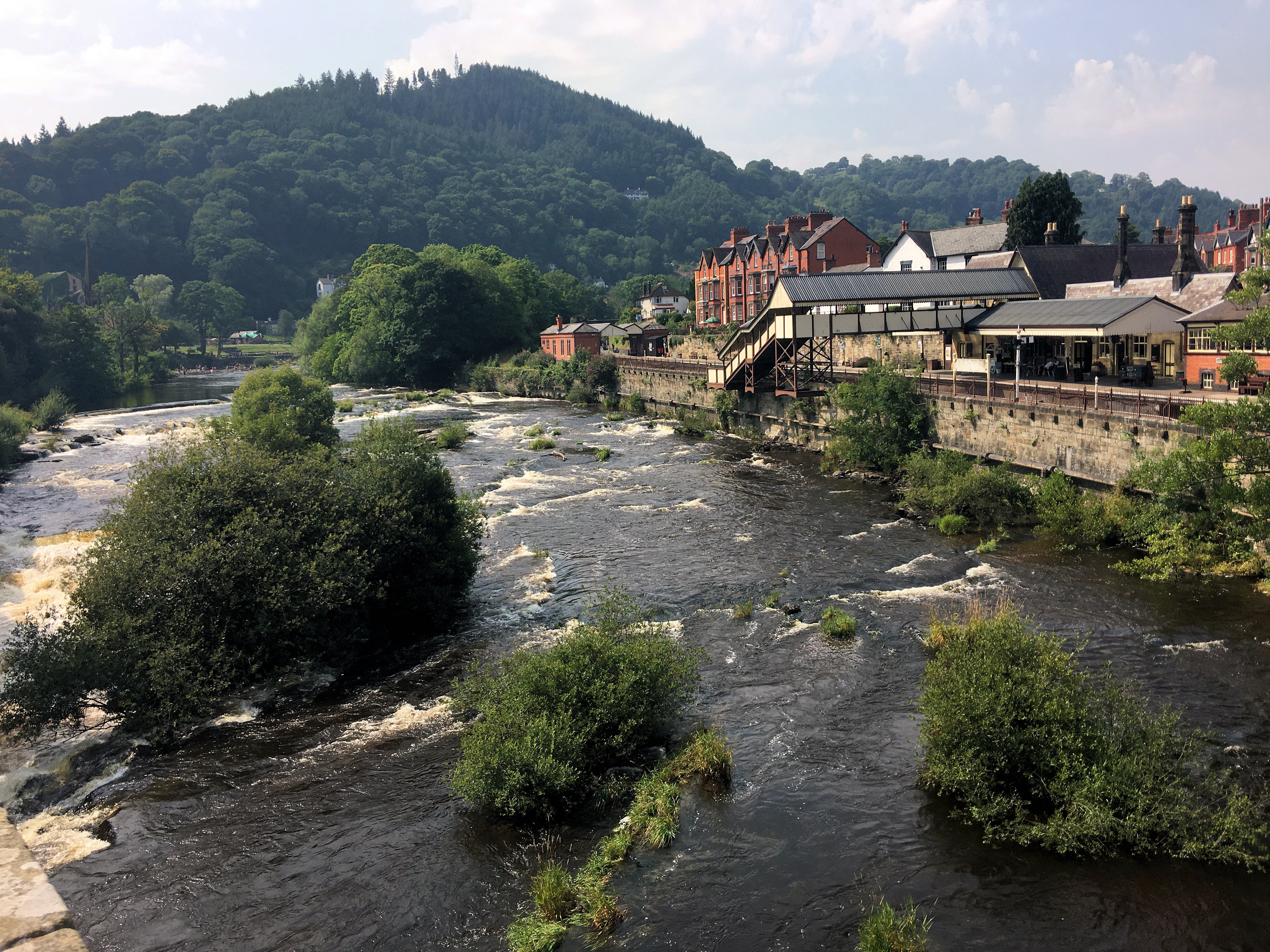 A scenic view of a river flowing through a town in Powys, with lush green hills in the background and a stone building along the riverside.