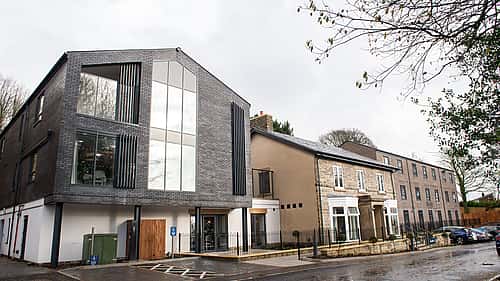 A modern building with sleek glass features stands next to a traditional stone structure, blending contemporary design with classic architecture.