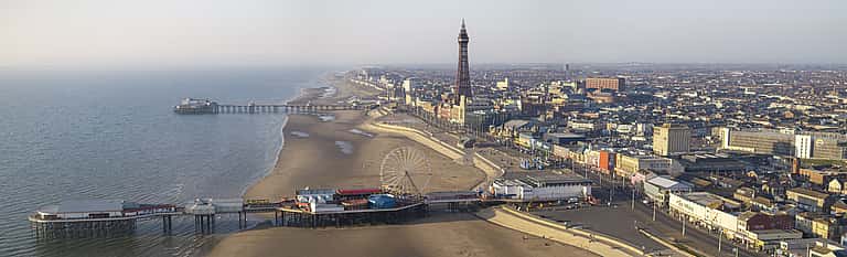 A panoramic view of Blackpool featuring its sandy beach, iconic piers, Ferris wheel, and the prominent Blackpool Tower rising above the seaside town.