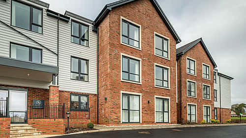 A modern brick and metal apartment building with large windows and a paved entrance area.