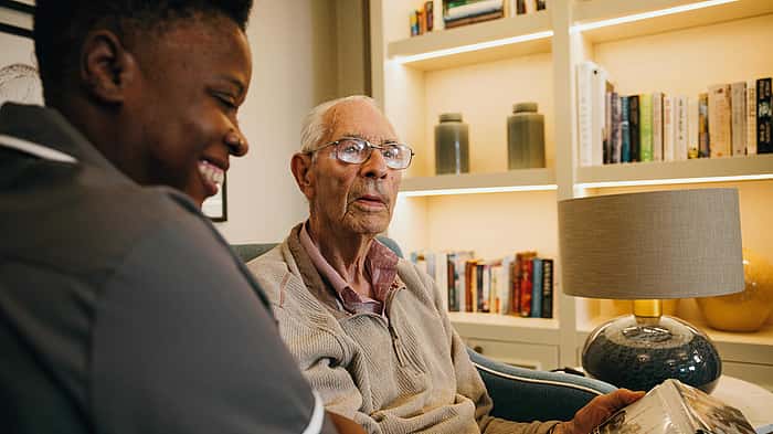 A caregiver and an elderly man smiling while looking at a photo album, surrounded by bookshelves and soft lighting