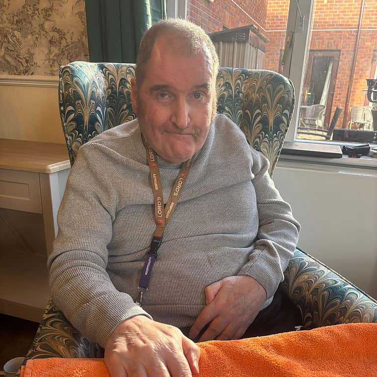 An older man sits comfortably with one hand resting on an orange towel, appearing calm and content during a pamper-style session.