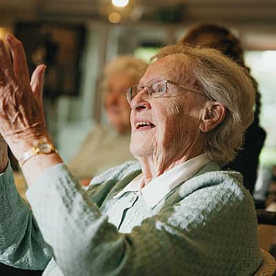 An elderly woman, dressed in a light blue sweater, joyfully claps her hands while sitting in a well-lit room