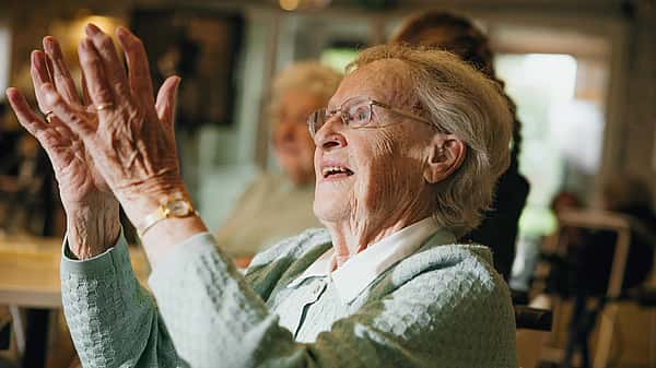 An elderly woman, dressed in a light blue sweater, joyfully claps her hands while sitting in a well-lit room