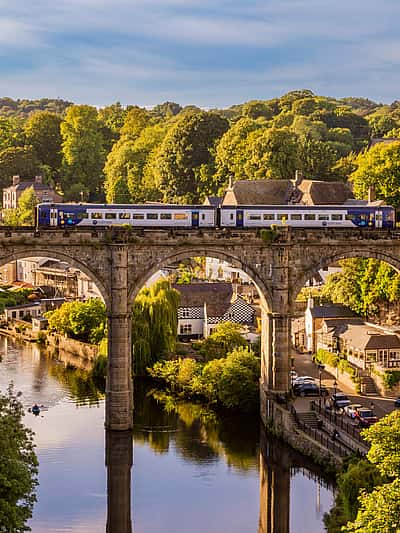 A scenic view of a train crossing a historic stone viaduct over a river, surrounded by lush greenery and charming buildings in North Yorkshire.