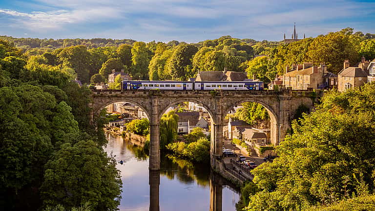 A scenic view of a train crossing a historic stone viaduct over a river, surrounded by lush greenery and charming buildings in North Yorkshire.