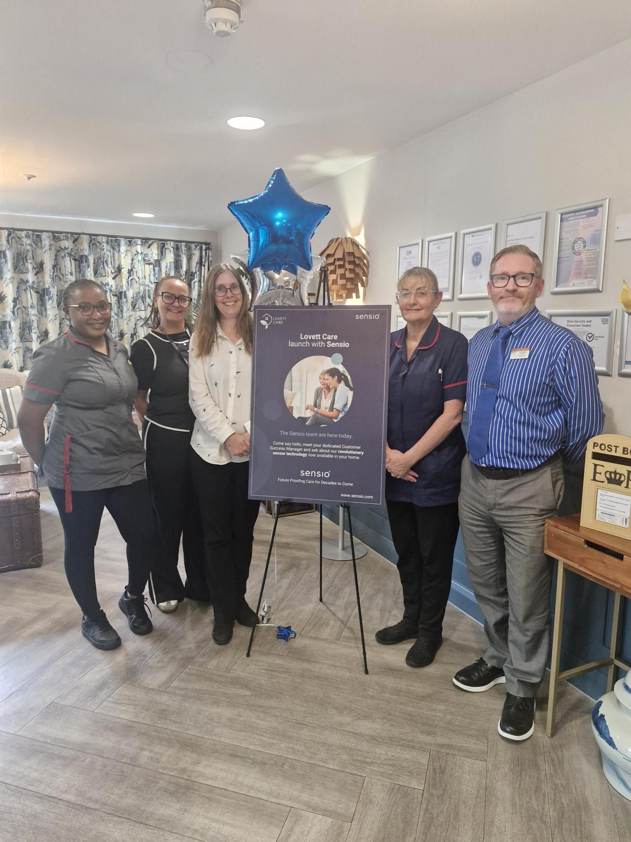 The image shows a group of six individuals standing together in a care home setting, with a large blue balloon and a poster in the background, likely celebrating a special event or achievement.