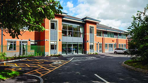 A modern two-story building with large windows, surrounded by a well-maintained parking area and greenery.