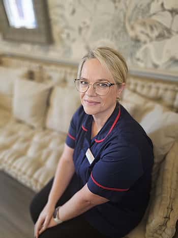 A lady in a purple top with blonde hair and glasses sat on a sofa.