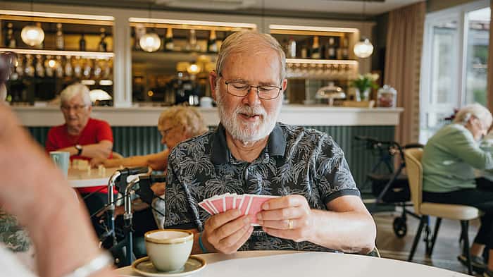An elderly man with glasses concentrates on his hand of cards while enjoying a cup of coffee in a cozy, well-lit café with others in the background
