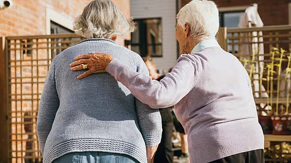 Two elderly women are walking together outdoors, with one gently placing her hand on the other's shoulder