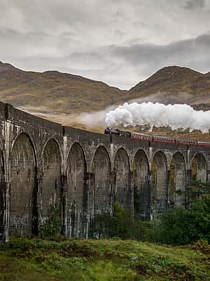 The image showcases a steam train crossing a stunning stone viaduct in a picturesque, mountainous landscape, possibly near Carlisle.