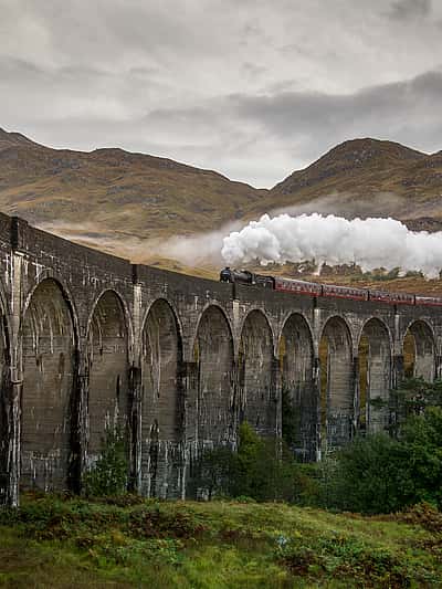 The image showcases a steam train crossing a stunning stone viaduct in a picturesque, mountainous landscape, possibly near Carlisle.
