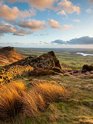 A scenic landscape featuring rolling hills in Staffordshire, a stone wall, golden grass, and a distant view of a river or lake under a partly cloudy sky at sunset.