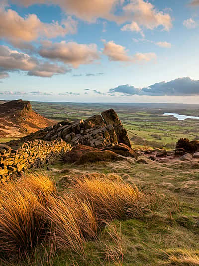 A scenic landscape featuring rolling hills in Staffordshire, a stone wall, golden grass, and a distant view of a river or lake under a partly cloudy sky at sunset.