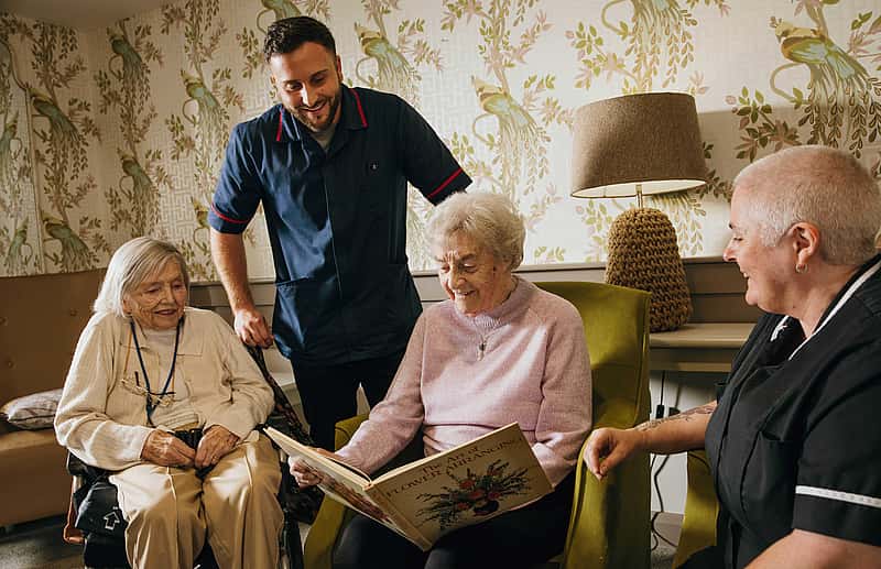 A group of elderly woman viewing a book. Two care professionals giving them company