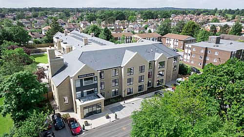 An aerial view of a modern residential building surrounded by greenery and houses, with cars parked along the street below.