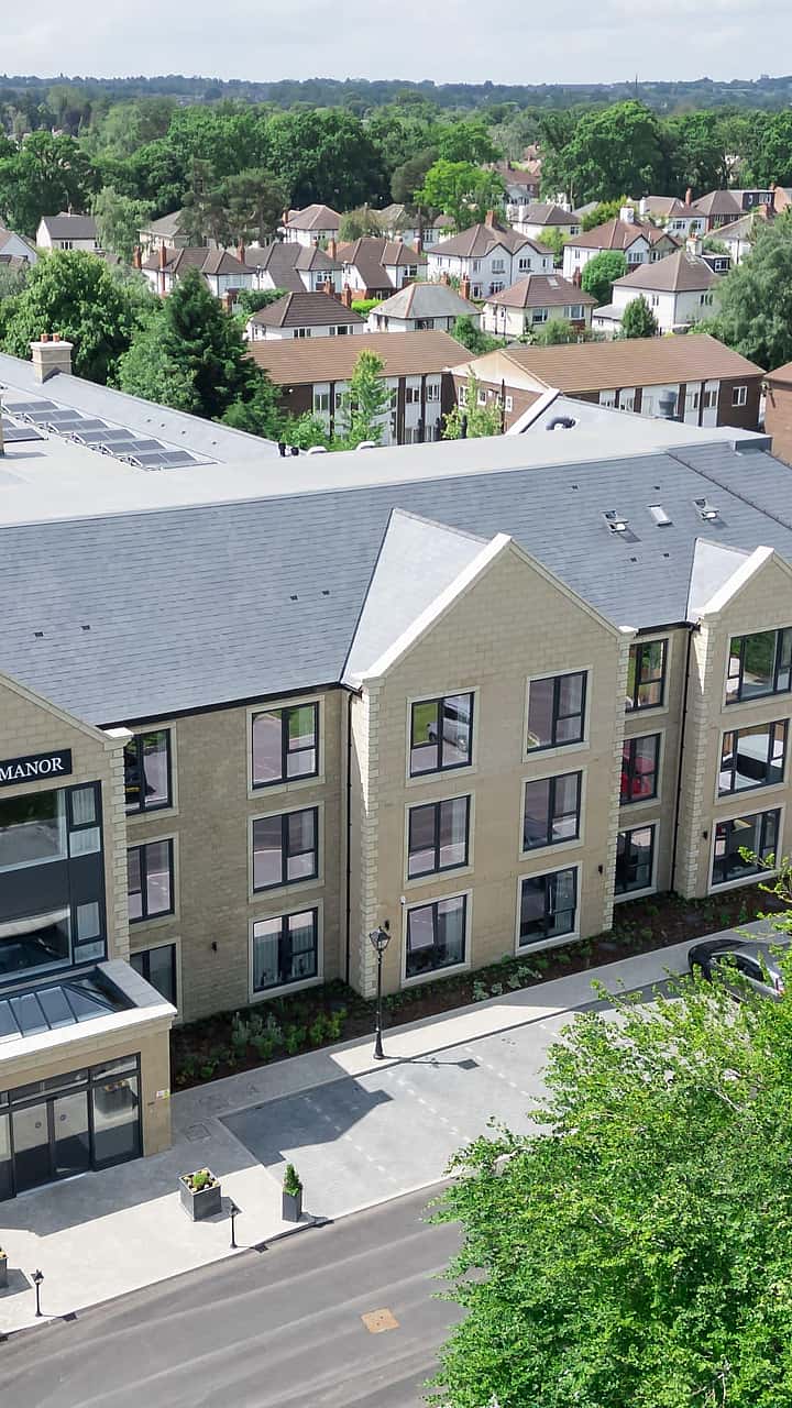 An aerial view of a modern residential building surrounded by greenery and houses, with cars parked along the street below.