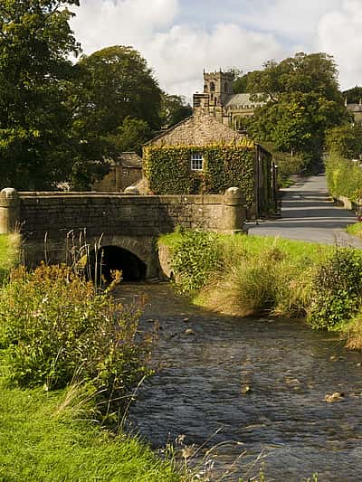 A scenic view of a quiet Lancashire village with a stone bridge over a stream, surrounded by lush greenery and traditional stone cottages.