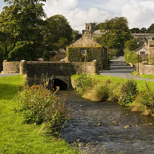 A scenic view of a quiet Lancashire village with a stone bridge over a stream, surrounded by lush greenery and traditional stone cottages.