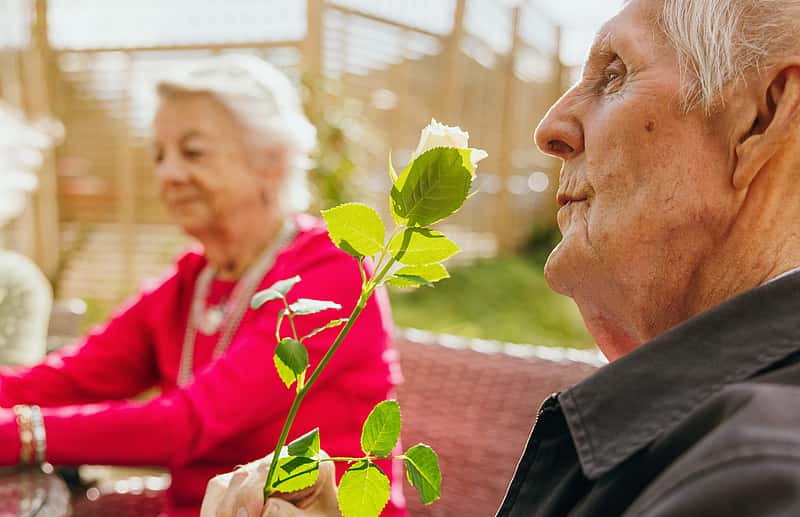 An elderly man holding a rose while a woman is seated in the background, both enjoying a peaceful moment outdoors