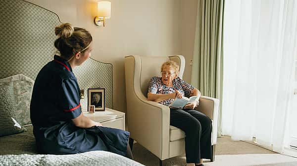 A caregiver in a dark uniform sits by a bed talking with an elderly woman reading a book in an armchair by a bright window