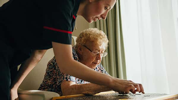 A caregiver helping an elderly woman with a puzzle on a table